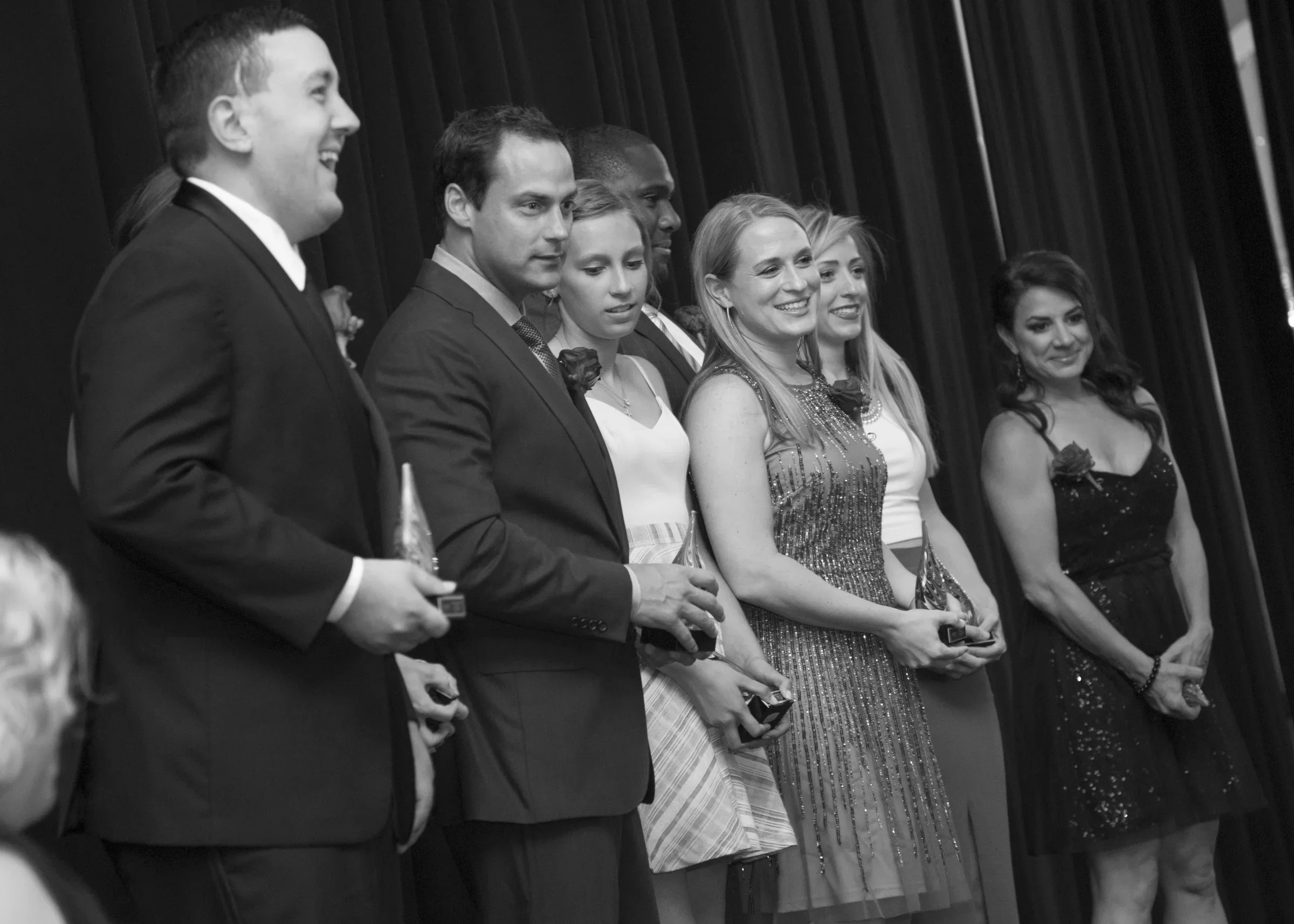 Group of people at an award ceremony holding trophies, dressed in formal attire, standing in front of a curtain.