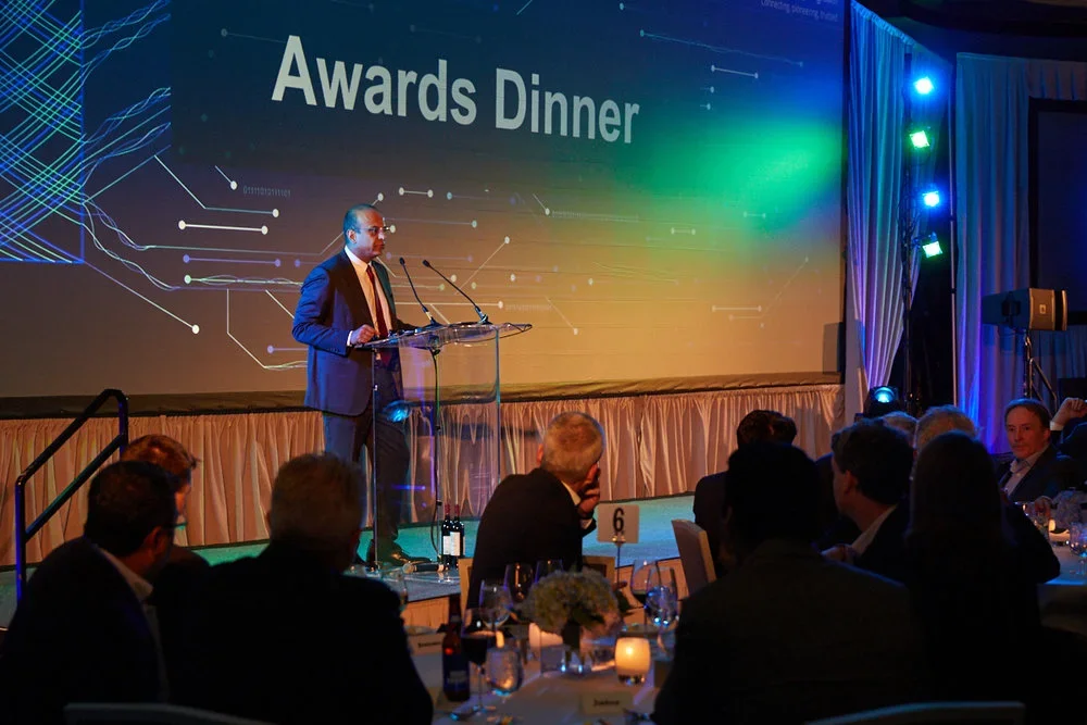 Man in dark suit speaking at a podium during an awards dinner, with a large screen displaying 'Awards Dinner' behind him, and seated guests at tables in the foreground.