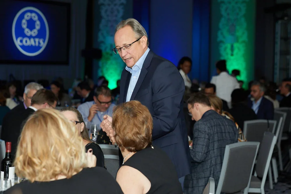 A man in a dark suit and glasses speaking to two women at a formal event with many other attendees at tables, with a screen showing a logo that says 'Coats' in the background.