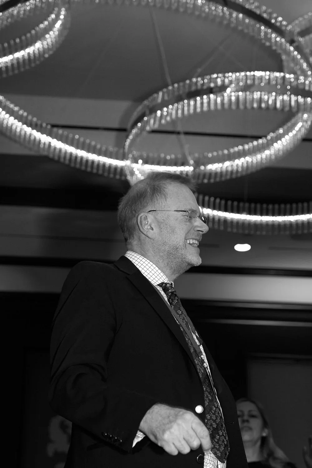 A smiling man in a suit and tie, wearing glasses, standing under a modern circular chandelier with hanging lights at an indoor event.