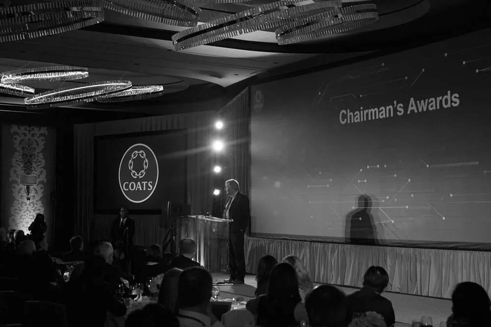 A black and white photo of an awards ceremony, featuring a stage with a large screen displaying "Chairman's Awards" and the logo of COATS. A person stands at the podium, with several attendees seated facing the stage.
