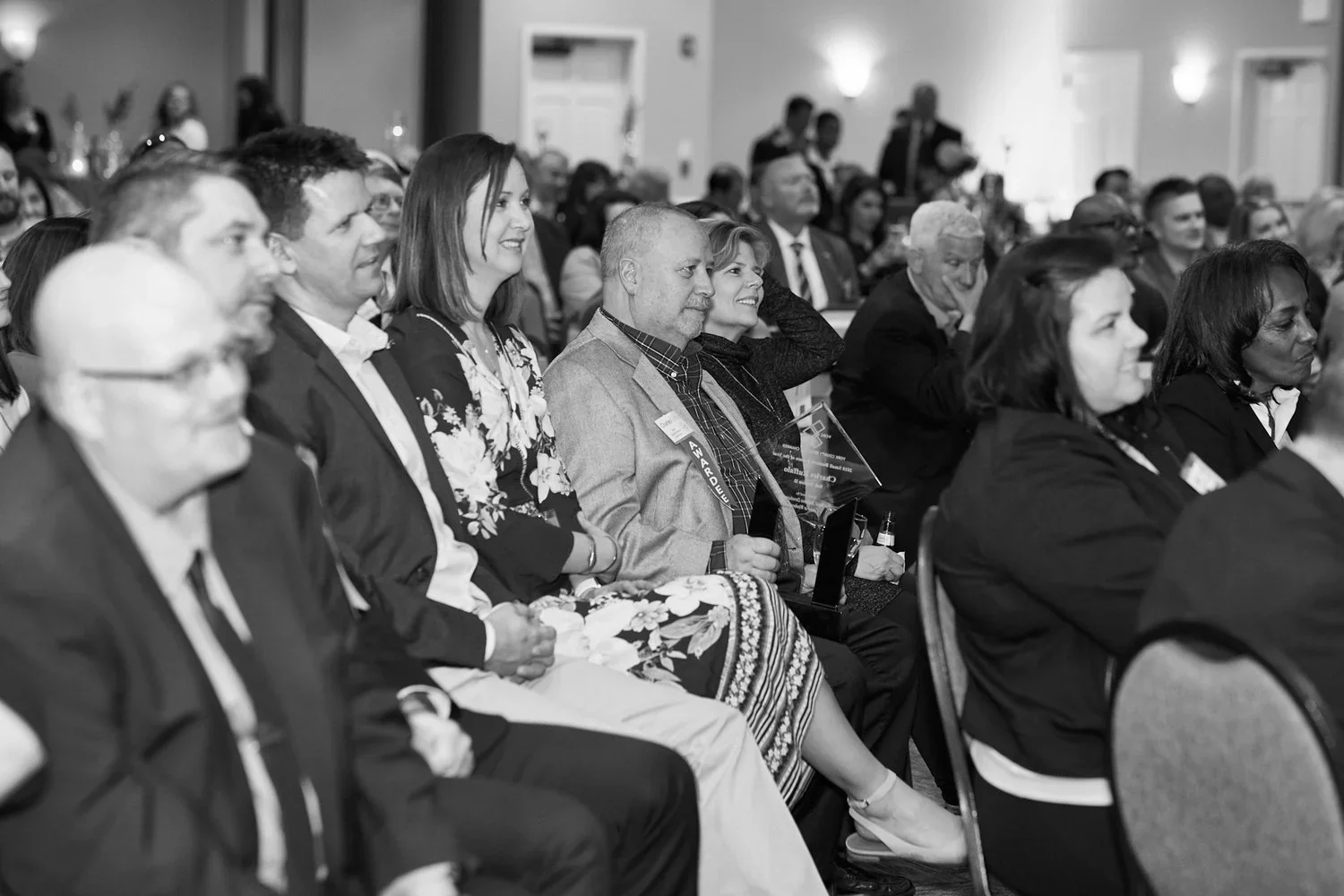Black and white photo of a group of people sitting in an audience at a formal event, some holding awards, with a few standing and mingling in the background.