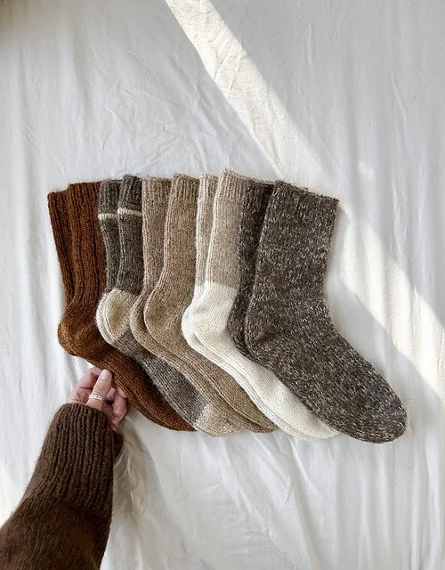 Six pairs of knitted socks in brown, gray, beige, and dark gray, displayed on a white surface with a hand holding the edge of the first pair.