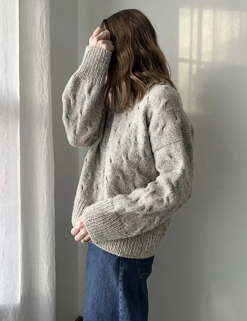 Side view of a woman with wavy brown hair wearing a beige knitted sweater and blue jeans, standing near a window with her hand touching her hair.