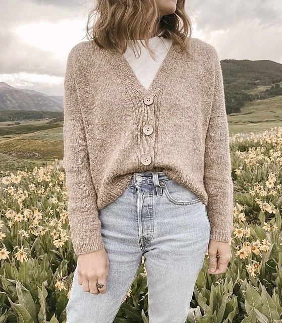 Woman wearing a beige cardigan with large buttons and light blue jeans standing in a field of yellow flowers with mountains in the background.