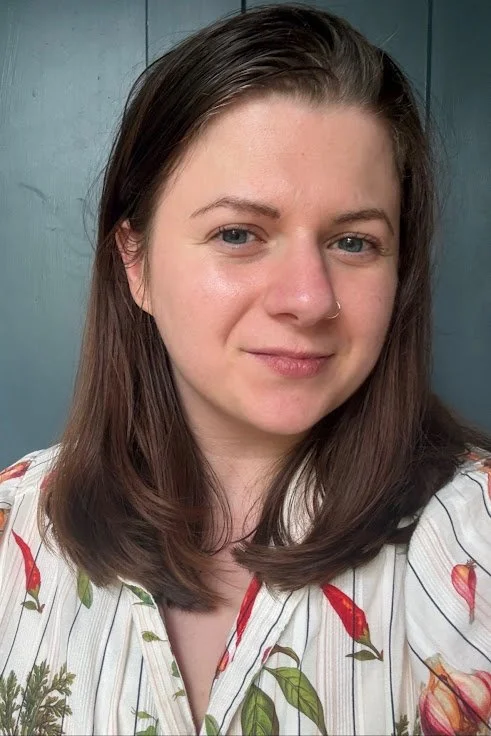 A young woman with shoulder-length brown hair, blue eyes, and a nose ring, smiling slightly. She is wearing a white shirt with a colorful floral and stripe pattern, and standing in front of a dark teal wooden wall.