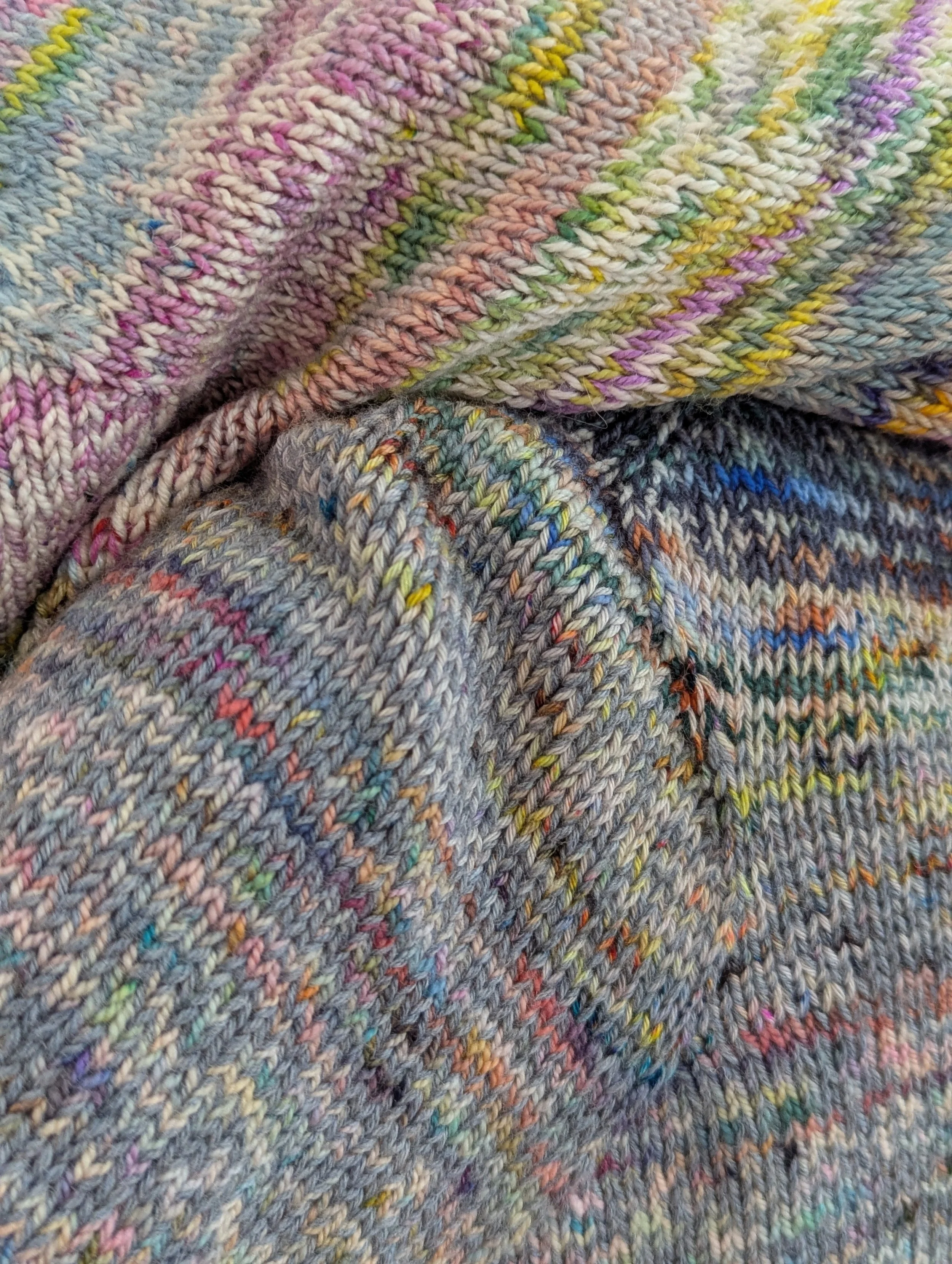 Close-up of multicolored knitted fabric with various shades of yarn, including pink, purple, yellow, green, blue, and gray, showing detailed knitting stitches.