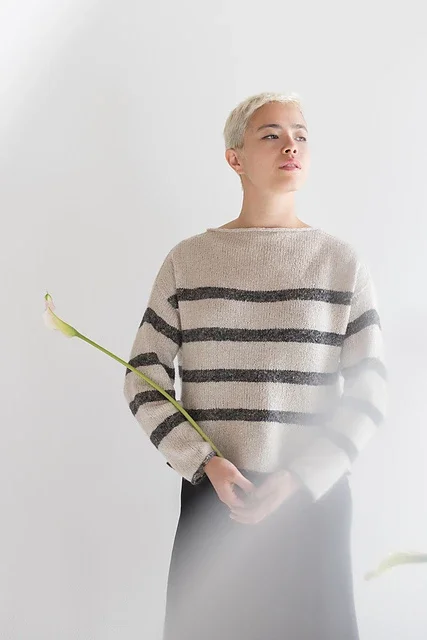 Person with short platinum blonde hair wearing a beige and black striped sweater, holding a single white calla lily flower.