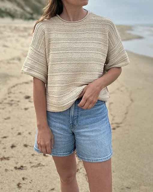 Person in a beige knit sweater and denim shorts standing on a sandy beach near the water.