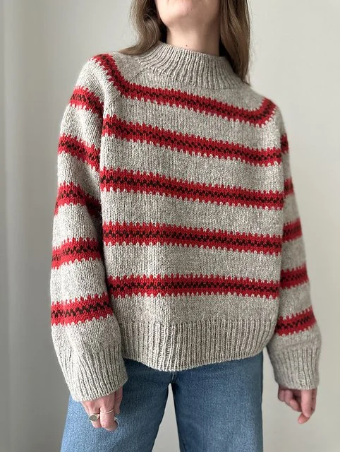 A person is wearing a beige knit sweater with red horizontal stripes and blue jeans, standing against a plain wall.