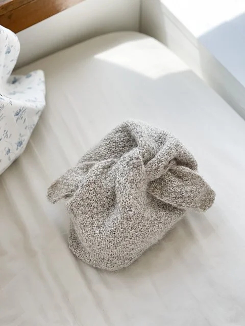 A cloth bag tied at the top, placed on a bed with white sheets and a floral pillow in the background.