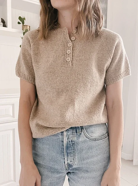 Woman wearing a beige knit top with buttons at the collar and light blue jeans, standing indoors in front of a white cabinet.