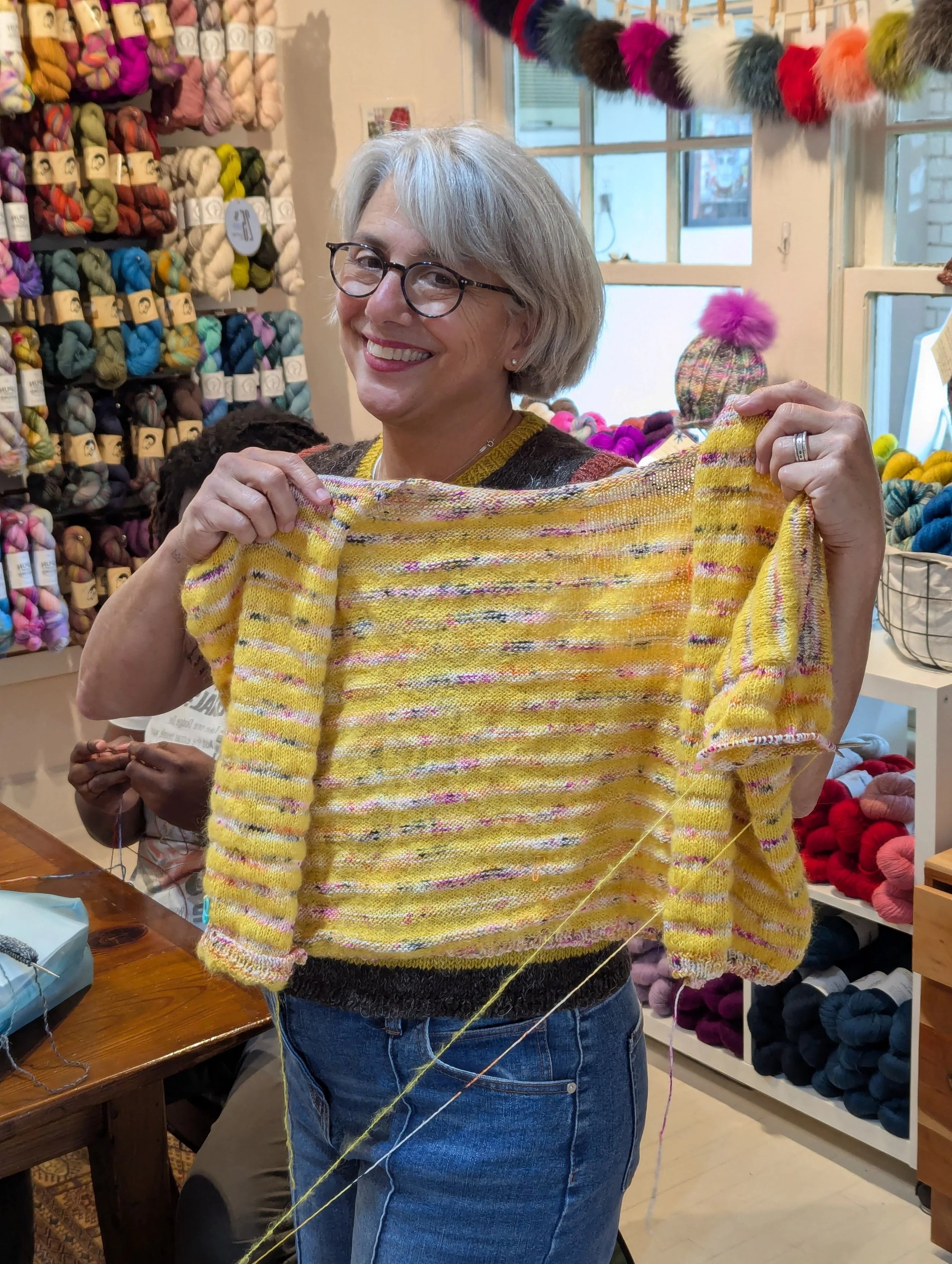 A woman with gray hair and glasses smiling, holding a yellow, pink, and purple striped yarn sweater in a yarn shop. Behind her, shelves display colorful yarns, and a window is visible with a colorful pom-pom decoration hanging inside.