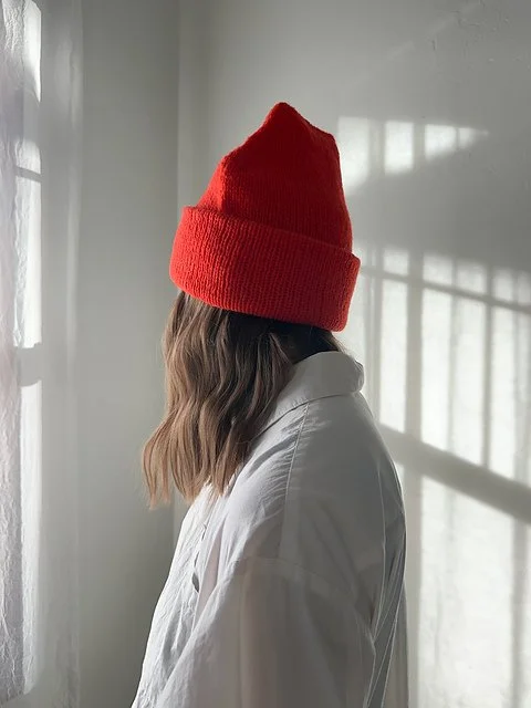 Person with brown hair wearing a red beanie hat and a white shirt, standing near a window with sunlight coming through.