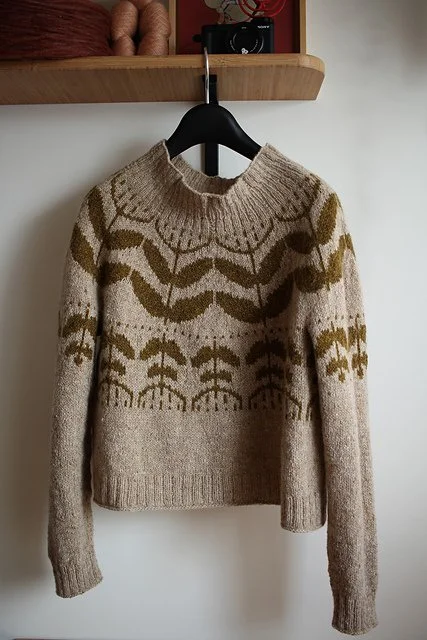 A beige sweater with brown and olive green leaf and branch patterns hanging on a black hanger against a white wall.