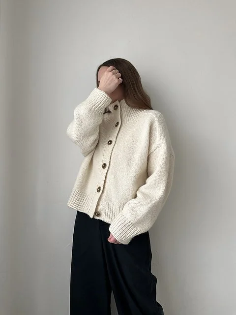 Person with long hair wearing a cream-colored button-up cardigan and black pants, standing against a plain white wall, covering part of their face with one hand.