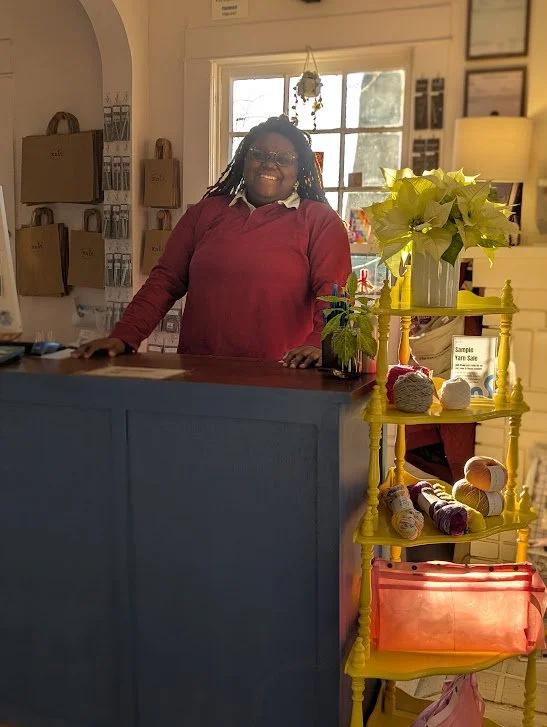 A woman with glasses and dreadlocks wearing a red sweater standing behind a navy blue counter in a cozy shop with various items on display, including a yellow shelf with yarn and decorative plants, and a window letting in natural light.