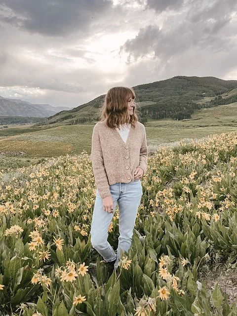Woman standing in a field of yellow flowers with mountains and cloudy sky in the background.