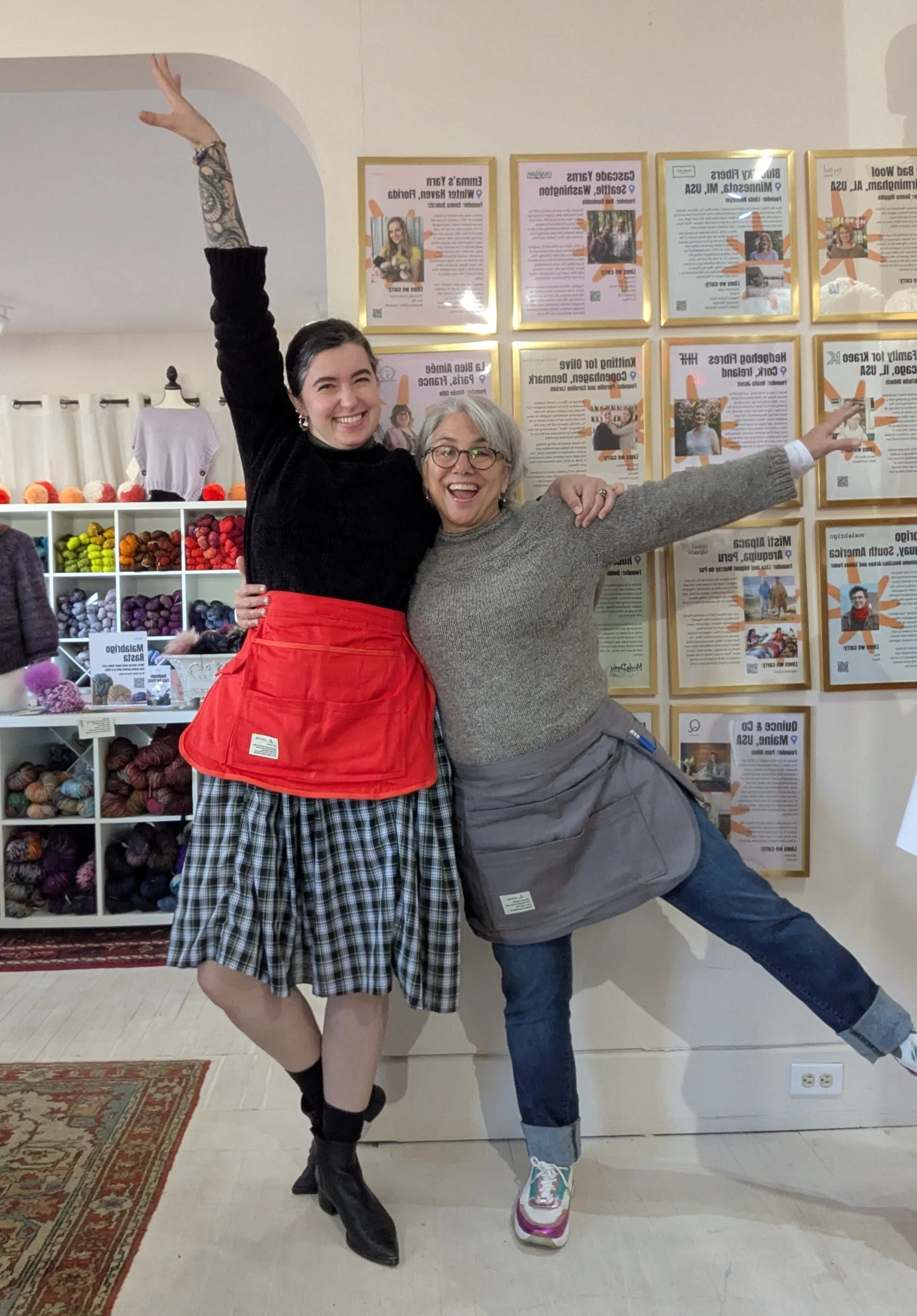 Two women smiling and posing happily in front of a wall of framed posters and a colorful yarn display.