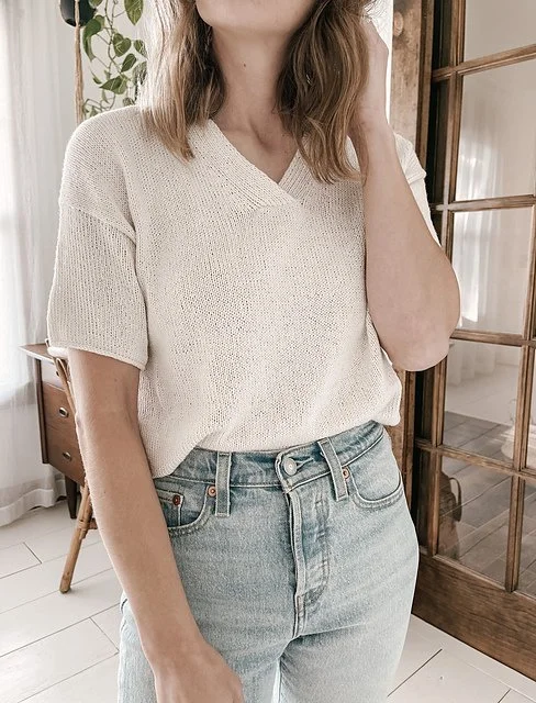 A woman wearing a cream-colored knit top and light blue jeans standing in a well-lit room with wooden and glass elements.