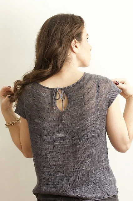 Back view of a woman with brown hair, wearing a gray Top, holding her hair with one hand, and wearing a gold bracelet.