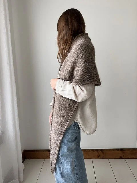 A person with long brown hair wearing a gray knitted shawl over a cream-colored shirt and blue jeans, standing indoors against a plain white wall.