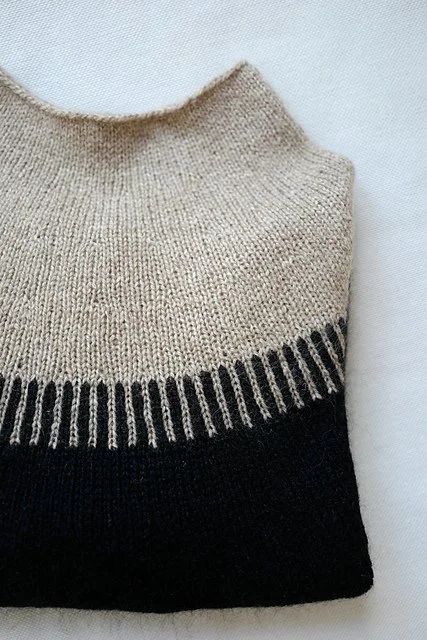 A beige sweater with brown and olive green leaf and branch patterns hanging on a black hanger against a white wall.