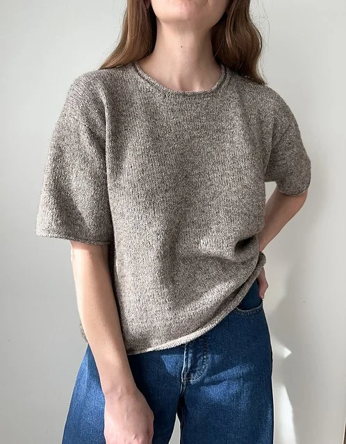 Person wearing a loose, short-sleeved gray sweater and blue jeans standing against a plain wall.