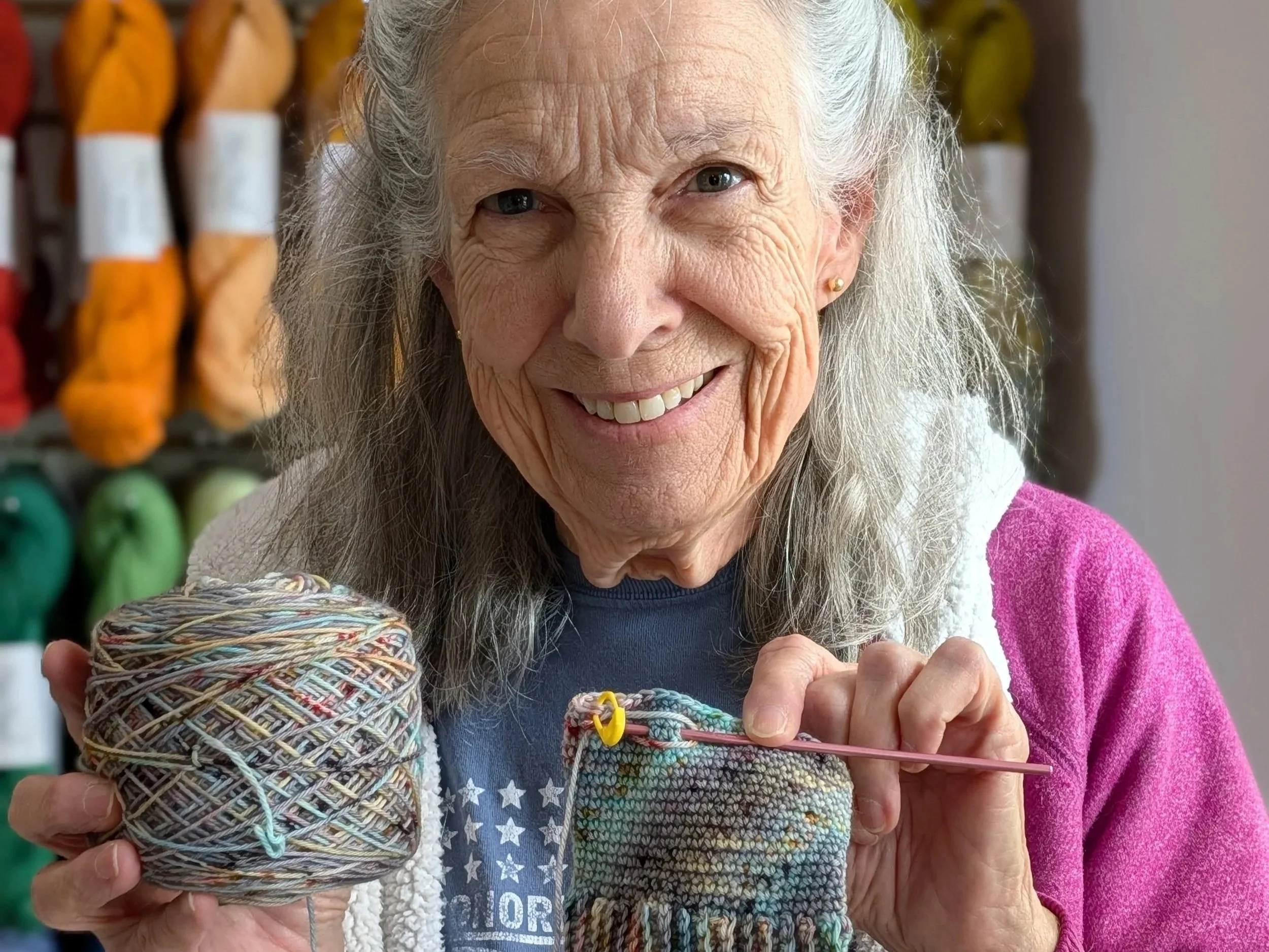 An elderly woman with white hair and earrings knitting with multicolored yarn in a cozy setting.