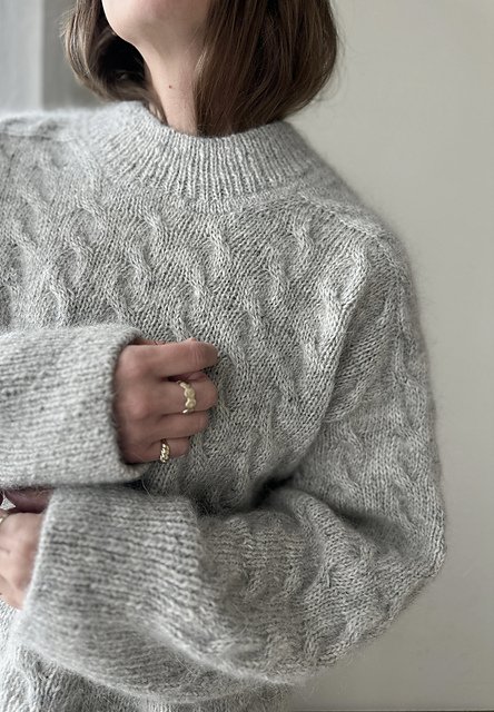 Close-up of a woman wearing a gray cable-knit sweater, with her hand resting on her chest, showing rings on her fingers.