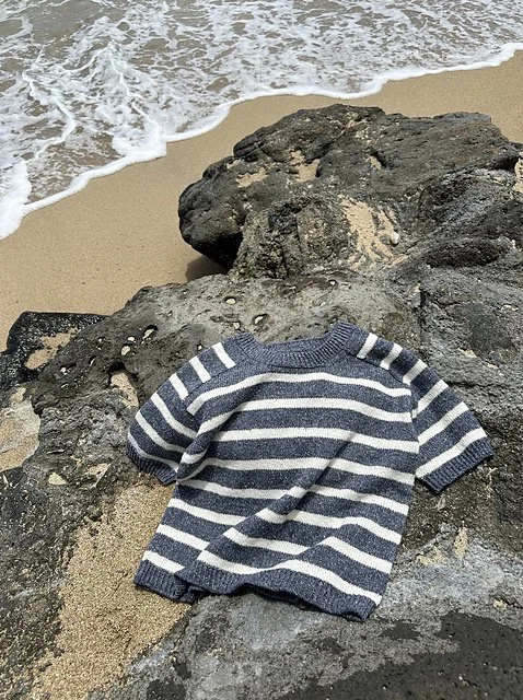 A striped navy and white sweater lying on a large dark rock at the beach with sand and ocean waves in the background.