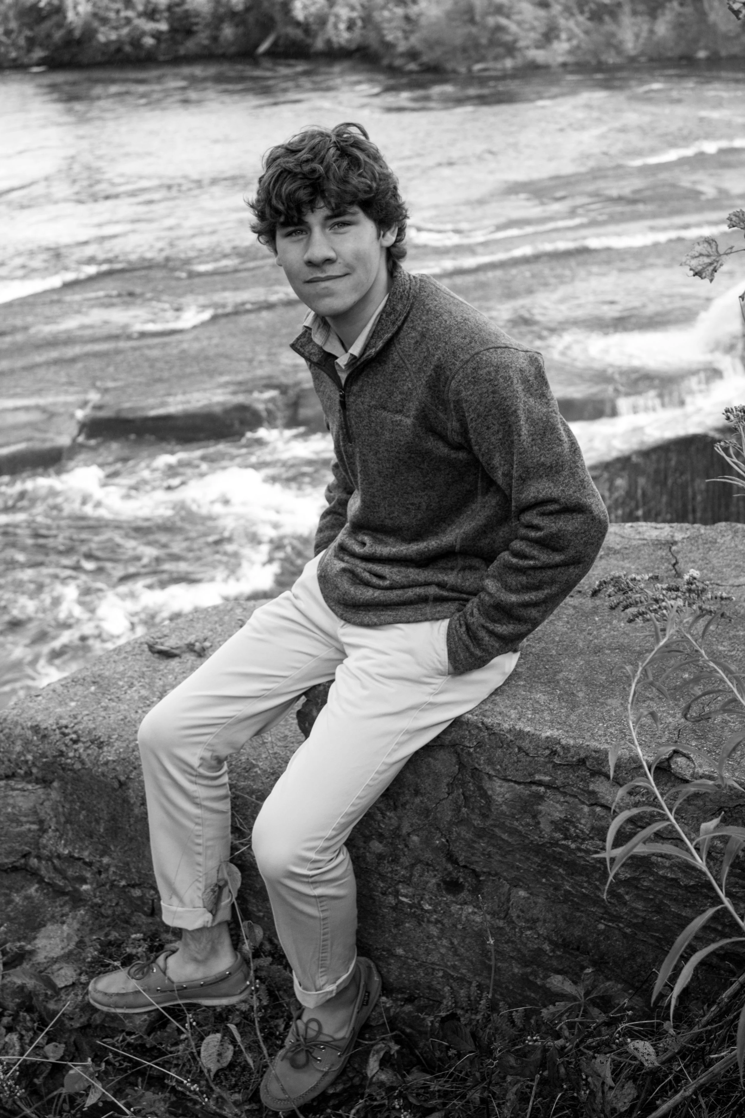 A young man with curly hair sitting on a rock near the shore of a body of water, wearing a zip-up sweater, light-colored pants, and boots, looking at the camera.