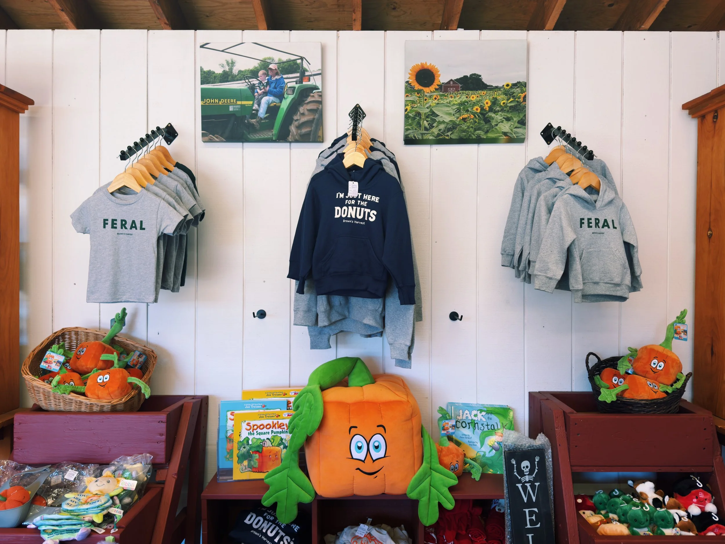 Display of farm-themed merchandise including T-shirts, hoodies, plush toys, and children's books with a harvest and sunflower theme, set against a white wall with framed farm landscape photographs.