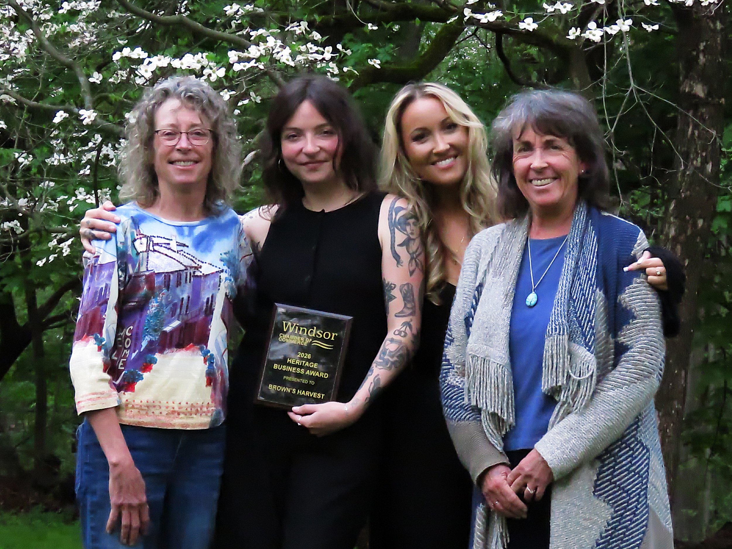 Four women standing outdoors in front of lush green trees, smiling at the camera. The second woman from the right is holding an award plaque that reads 'Windsor Chamber of Commerce 2026 Heritage Business Award presented to Brown's Harvest.'