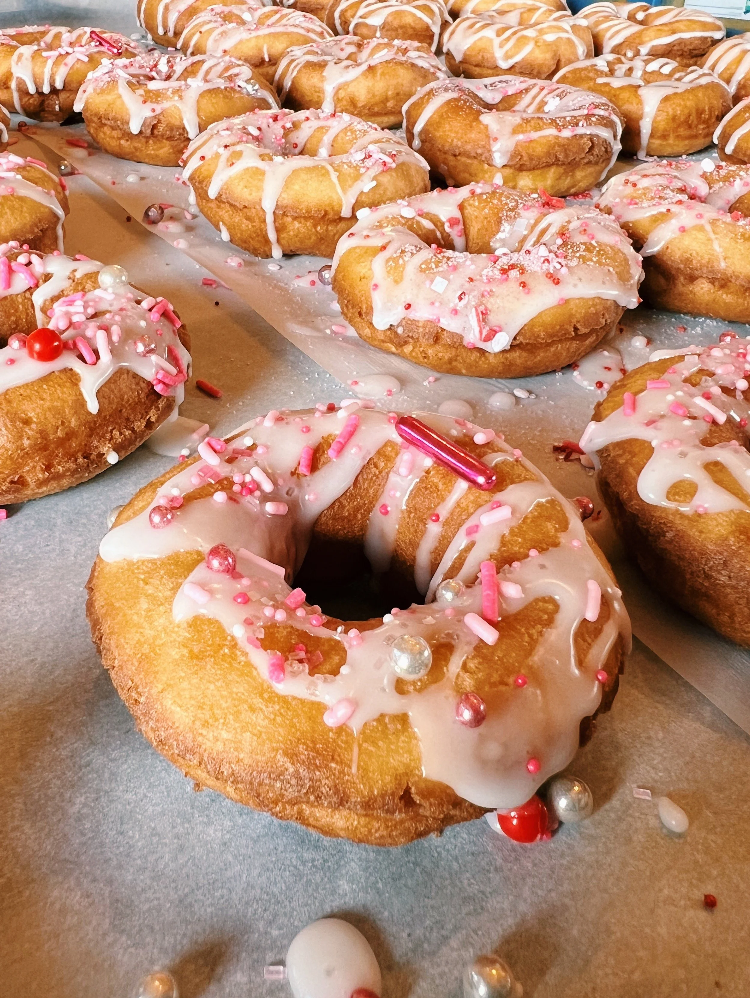 Close-up of assorted donuts topped with white icing, pink and white sprinkles, and decorative sugar pearls, placed on a baking sheet with scattered sprinkles and pearls around.
