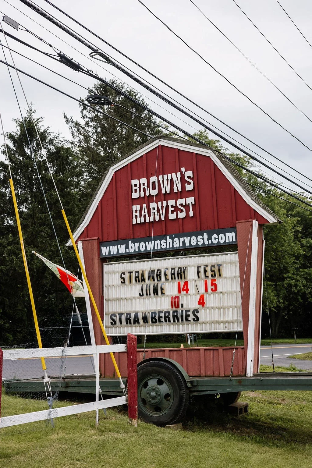 A red barn-style sign with white lettering advertising Brown's Harvest, a website link, and a schedule for a strawberry festival on June 14 and 15, with strawberries available on June 10 and 4.