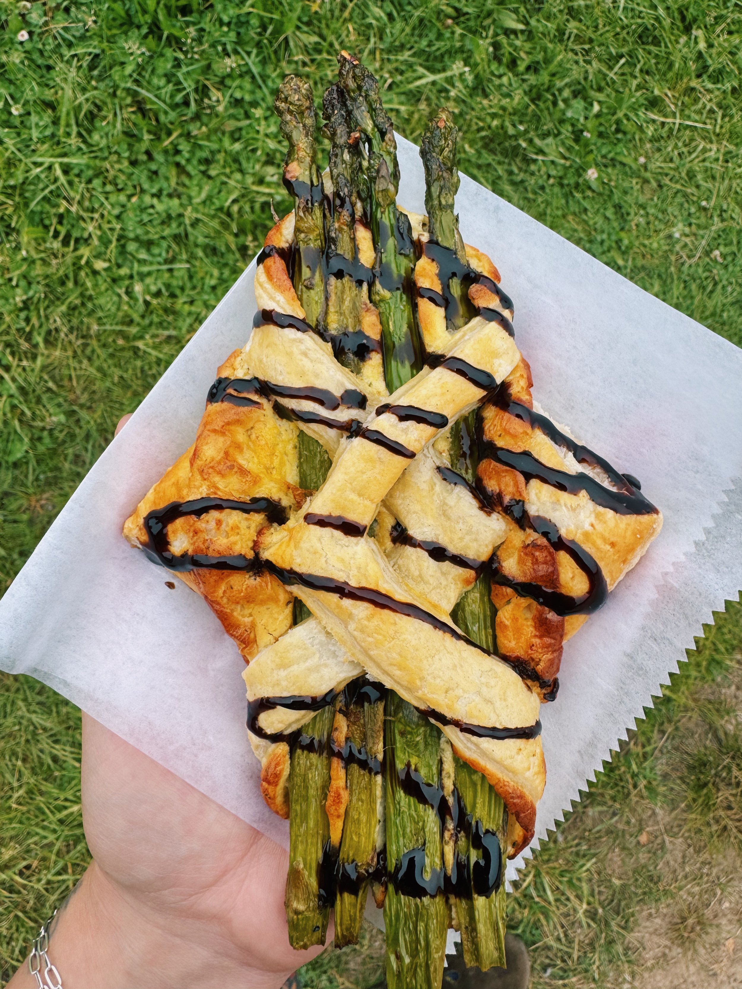 A person holding a grilled pizza topped with asparagus, drizzled with balsamic glaze, on a paper plate outdoors.
