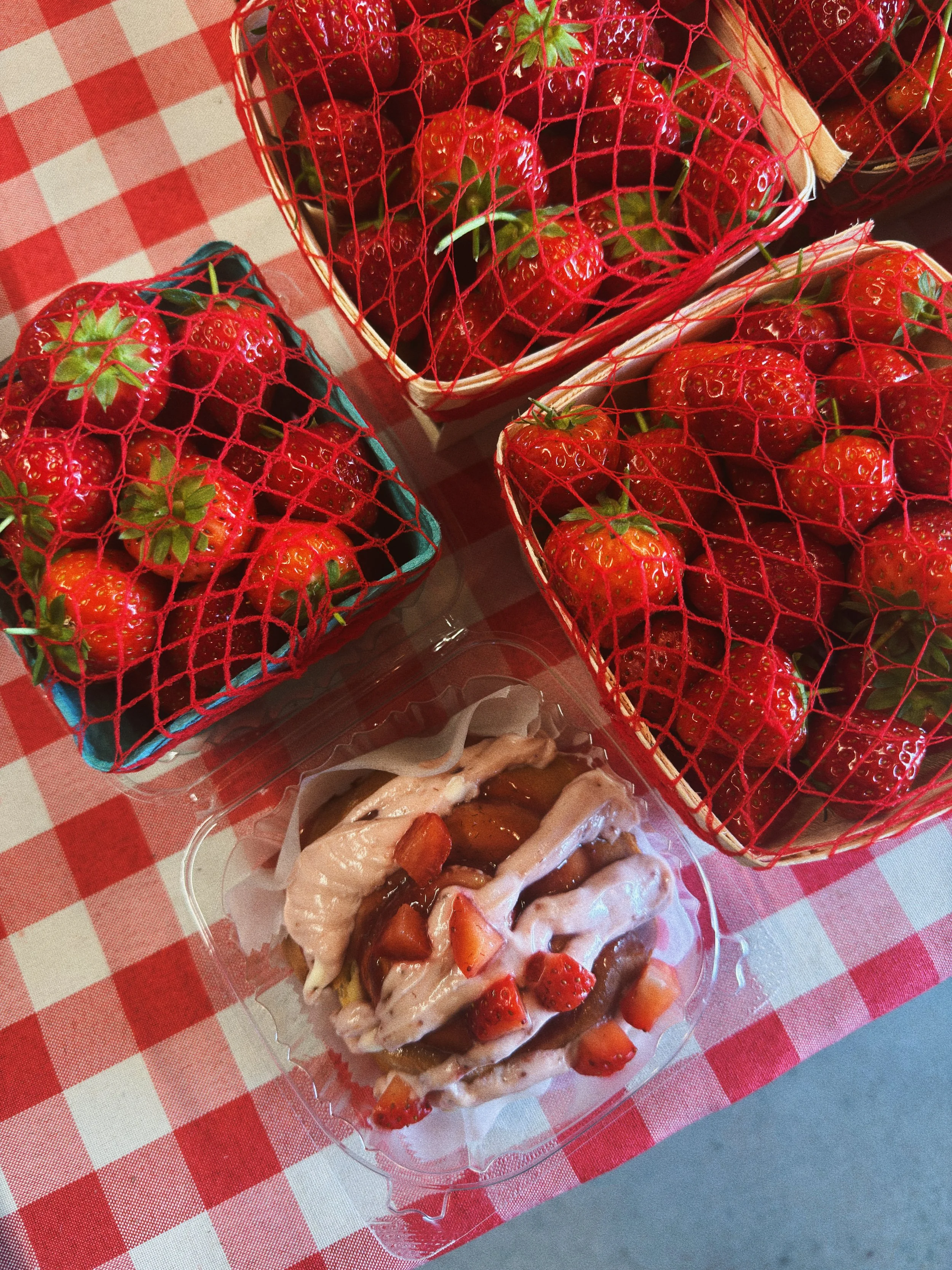 Baskets of fresh strawberries on a red and white checkered tablecloth, with a strawberry dessert topped with strawberries and syrup in a clear plastic container.