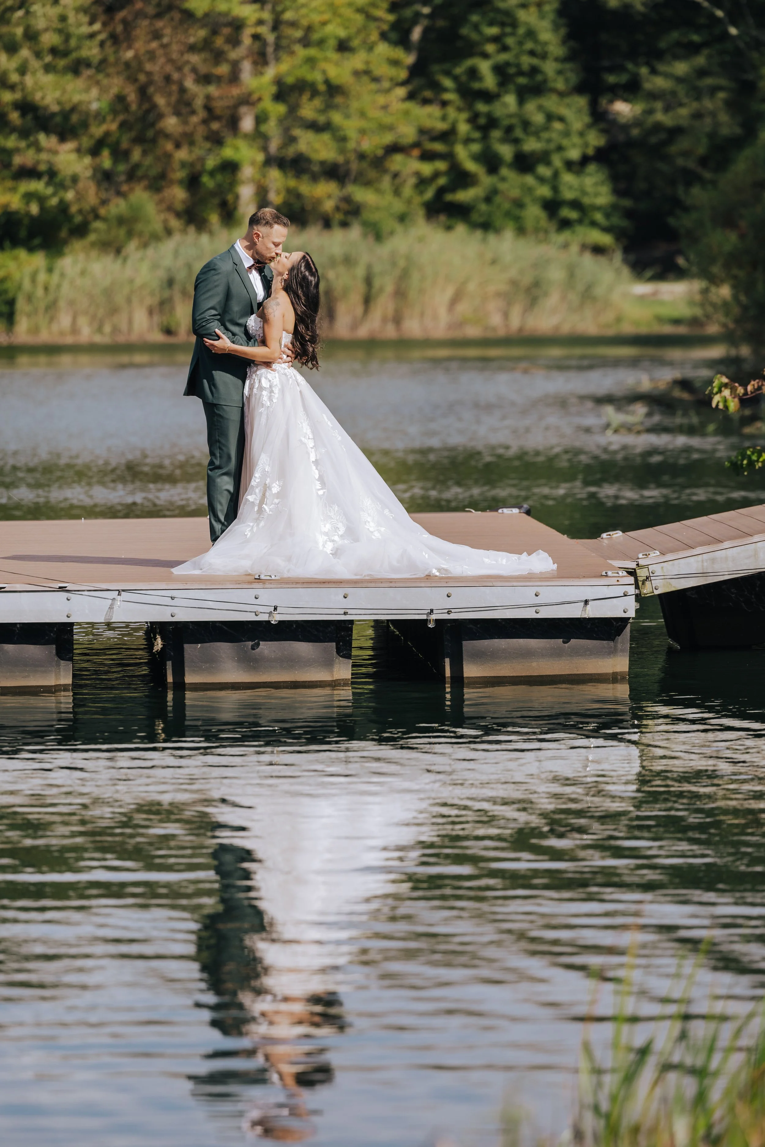 A bride and groom sharing a kiss on a wooden dock by a lake with lush green trees in the background.