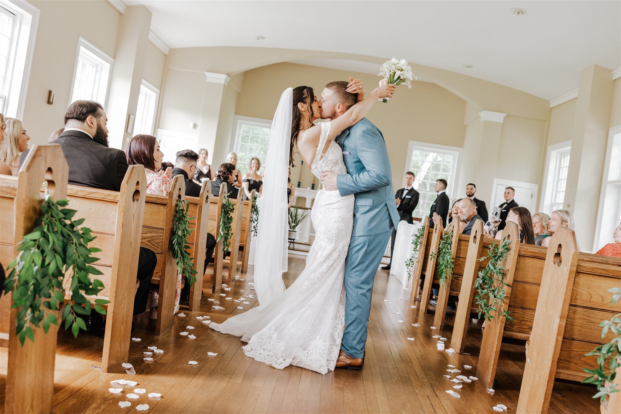 Bride and groom kissing during their wedding ceremony inside a decorated church, with guests watching and smiling.