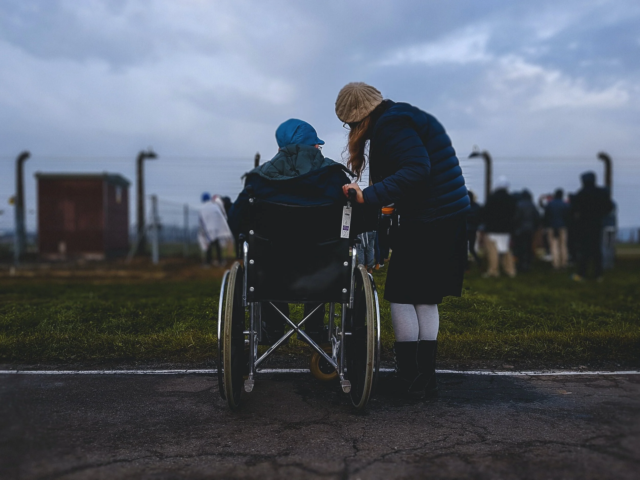 A person giving assistance to a person in a wheelchair outdoors during cloudy weather, with a group of people blurred in the background.