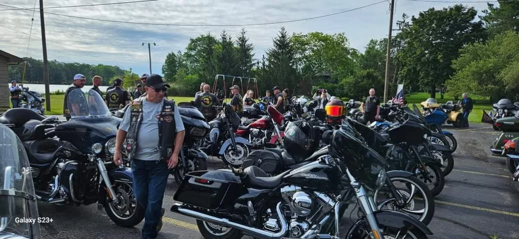 A group of motorcyclists gathered in a parking lot, with many bikes parked and some people standing and walking around, under cloudy skies and greenery in the background.
