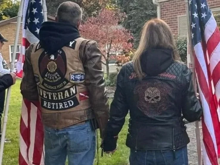 Two individuals, a man and a woman, stand side by side holding hands in front of American flags. The man wears a brown vest with patches that read "Veteran Retired" and a retirement badge, while the woman wears a black jacket with a skull design on t