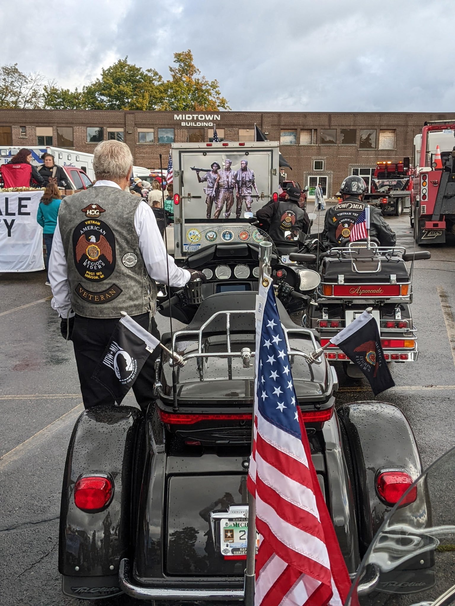 A motorcycle and a motorcycle sidecar with American flags and veterans patches, parked outside a building with a crowd and a large photo of soldiers in the background.