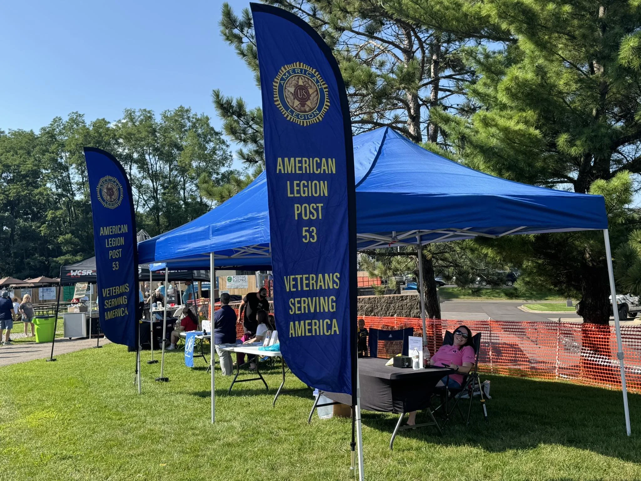 A blue canopy tent with two large blue flags displaying the American Legion Post 53 emblem and the slogan "Veterans Serving America." Several people are seated and standing around the tent, which is set up on a grassy area with trees and a parking lot in the background on a sunny day.
