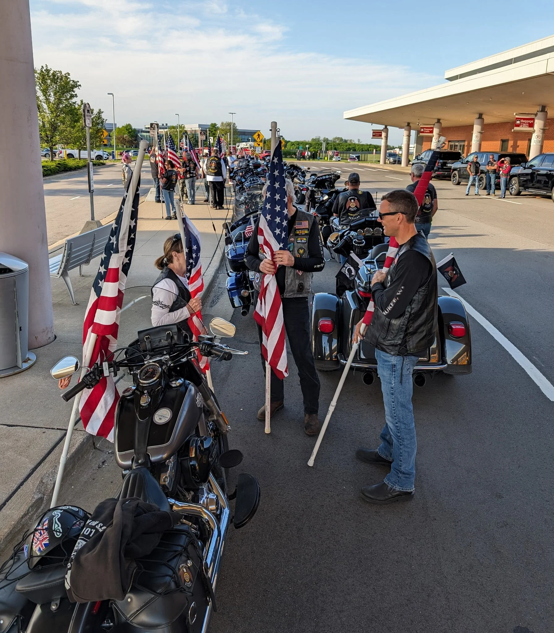 Group of motorcycle riders gathered outside a building holding American flags, with their bikes parked in line.