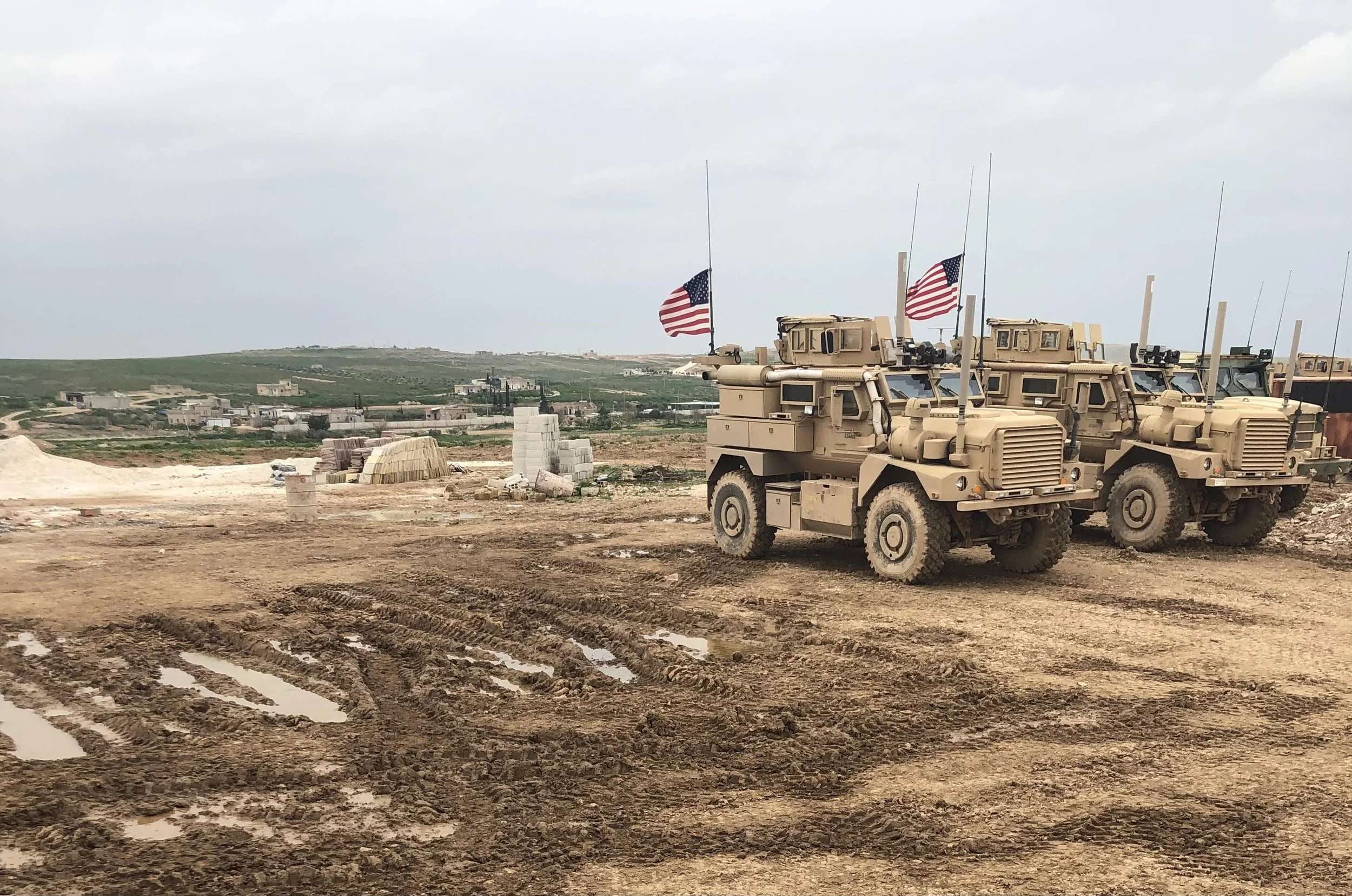 Three military vehicles with American flags parked on a muddy construction site, with a landscape of hills and scattered buildings in the background.