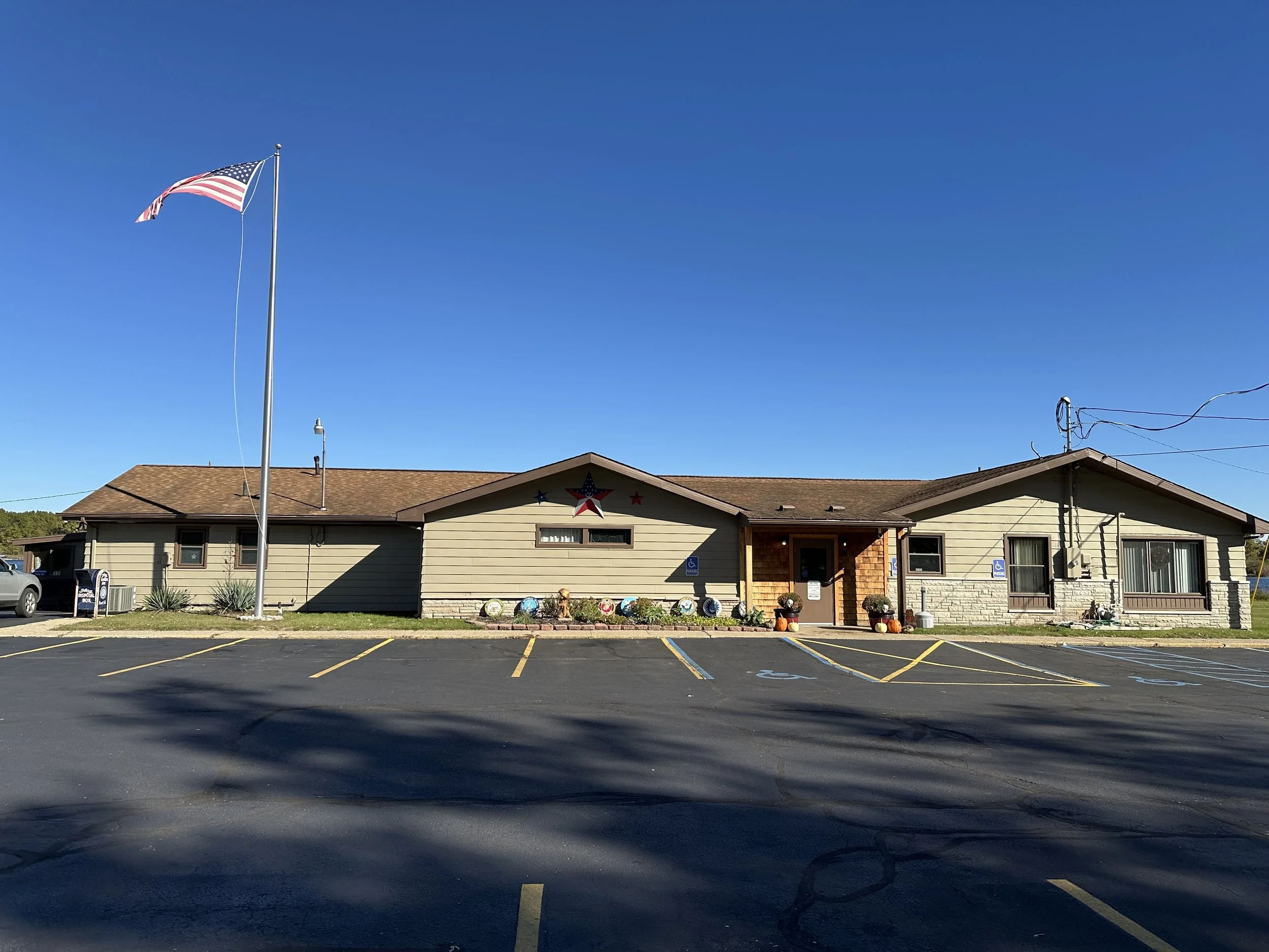 A single-story building with beige siding and a brown roof, decorated with patriotic stars and ornaments, with a parking lot in front and an American flag flying on a tall flagpole.