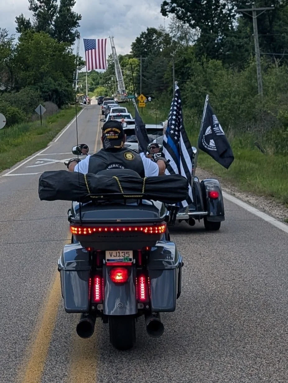 A motorcycle rider and a trailer with flags, including the American flag, on a rural road with line of parked cars and a toppled ladder in the background.