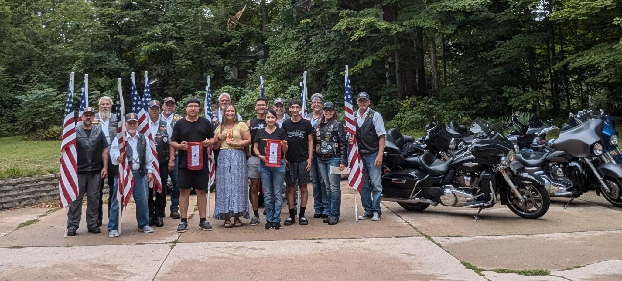 Group of people, some holding American flags, gathered outdoors with motorcycles parked on the right and a wooded background.